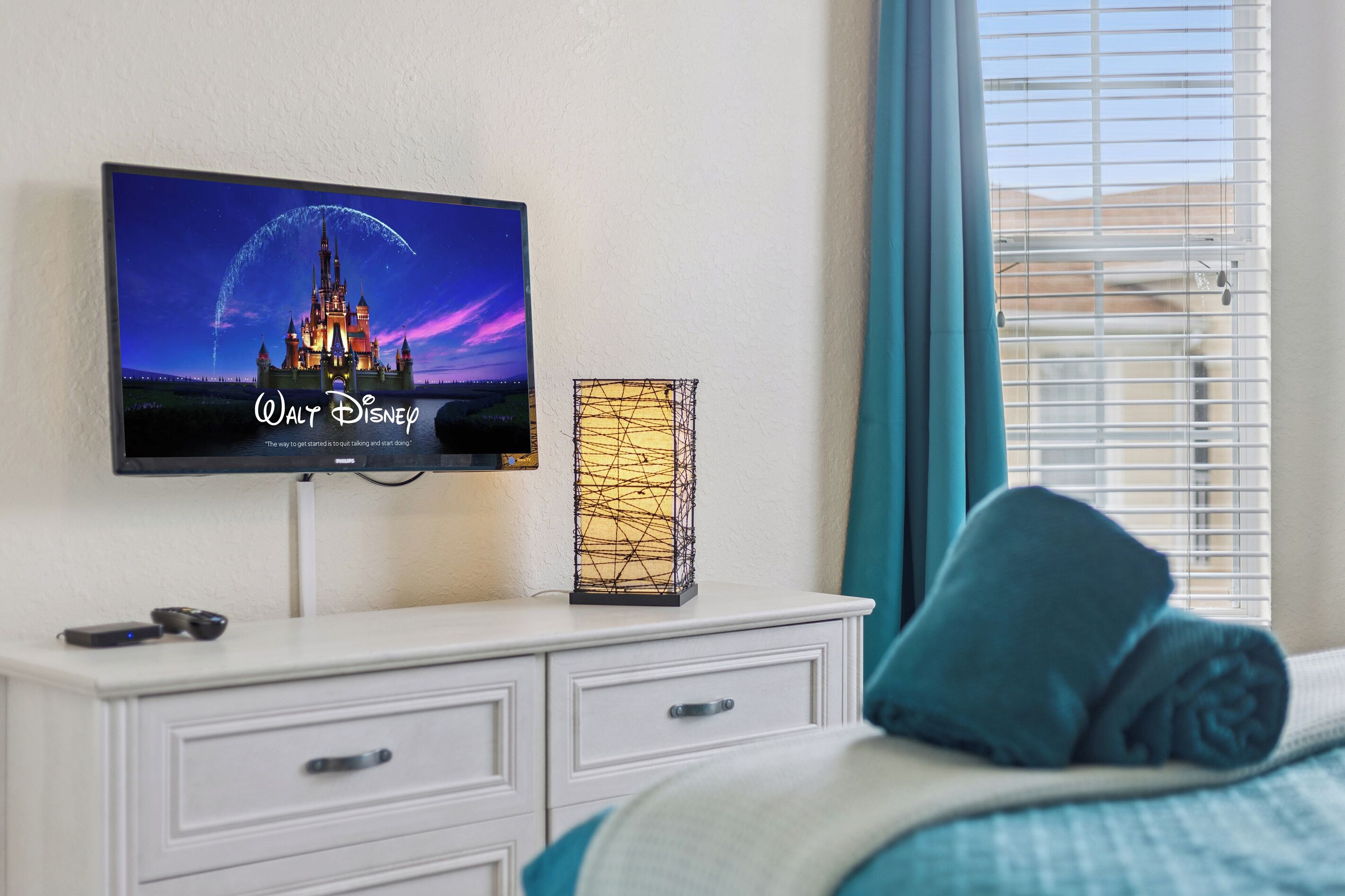 A television mounted on a wall displaying a castle, a white dresser with drawers, a decorative lamp, and a bed with a blue pillow.