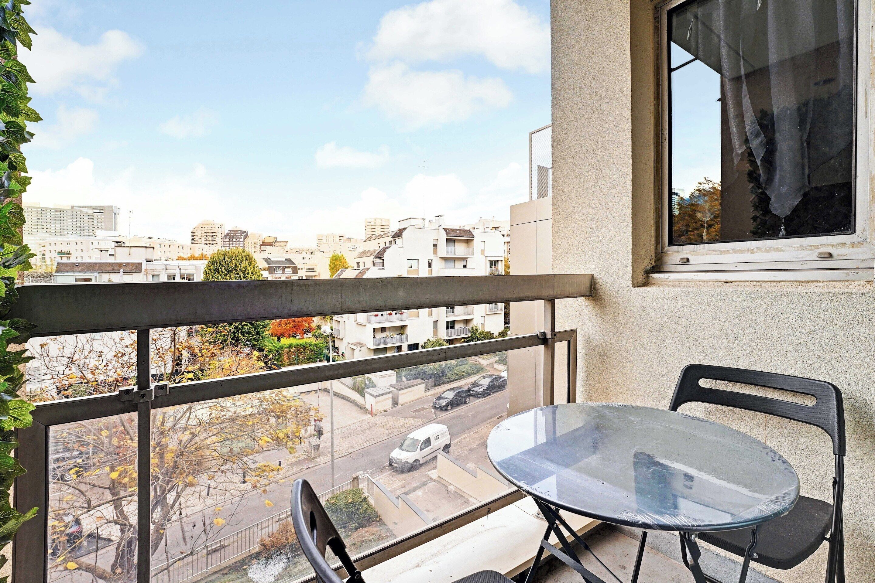 Uma varanda com mesa e cadeiras, vista para a paisagem urbana e uma janela com cortina.