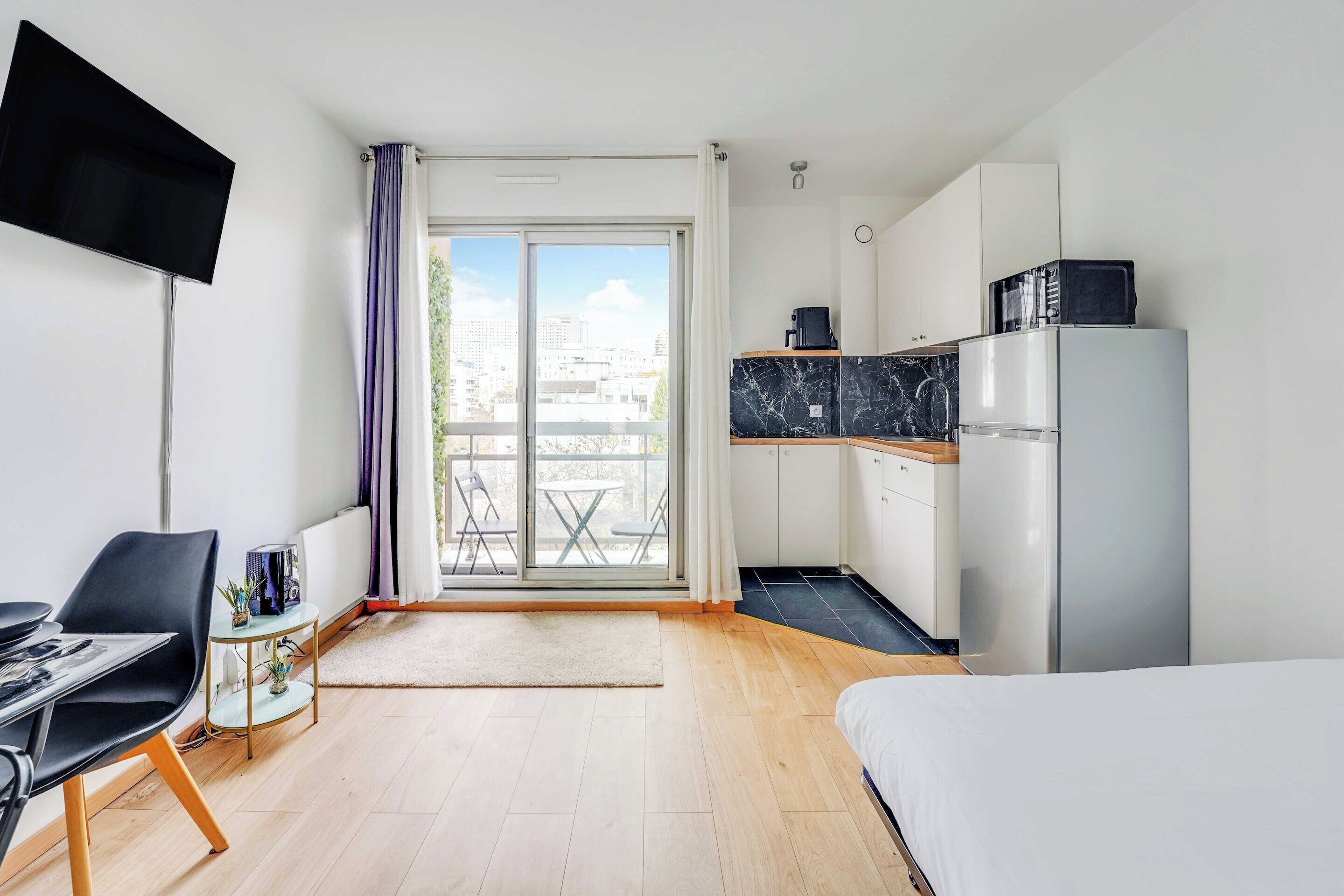 Une chambre d’hôtel moderne comprenant un lit, une table à manger avec des chaises, un petit coin cuisine et un balcon avec vue sur la ville.
