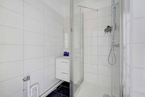 A modern bathroom with white tiles, a glass-enclosed shower, a white cabinet with drawers, and a black tiled floor. - Charming Studio 2P - Courbevoie (Courbevoie)