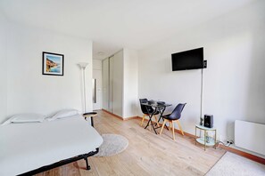 A bedroom with a bed, a desk, a chair, a TV, and a small table with a speaker. - Charming Studio 2P - Courbevoie (Courbevoie)
