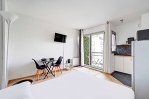 A modern room with a bed, a dining area with chairs, a TV, a kitchen area with a fridge, and a sliding glass door to the outside. - Charming Studio 2P - Courbevoie (Courbevoie)