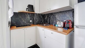 Une cuisine compacte avec un dosseret en marbre noir, des armoires blanches, un évier en acier inoxydable et une machine à expresso rouge.