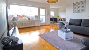 Ein Wohnzimmer mit grauem Sofa, einem Couchtisch mit Tablett, einem Fernseher auf einem Schrank und Blick ins Freie durch große Fenster.