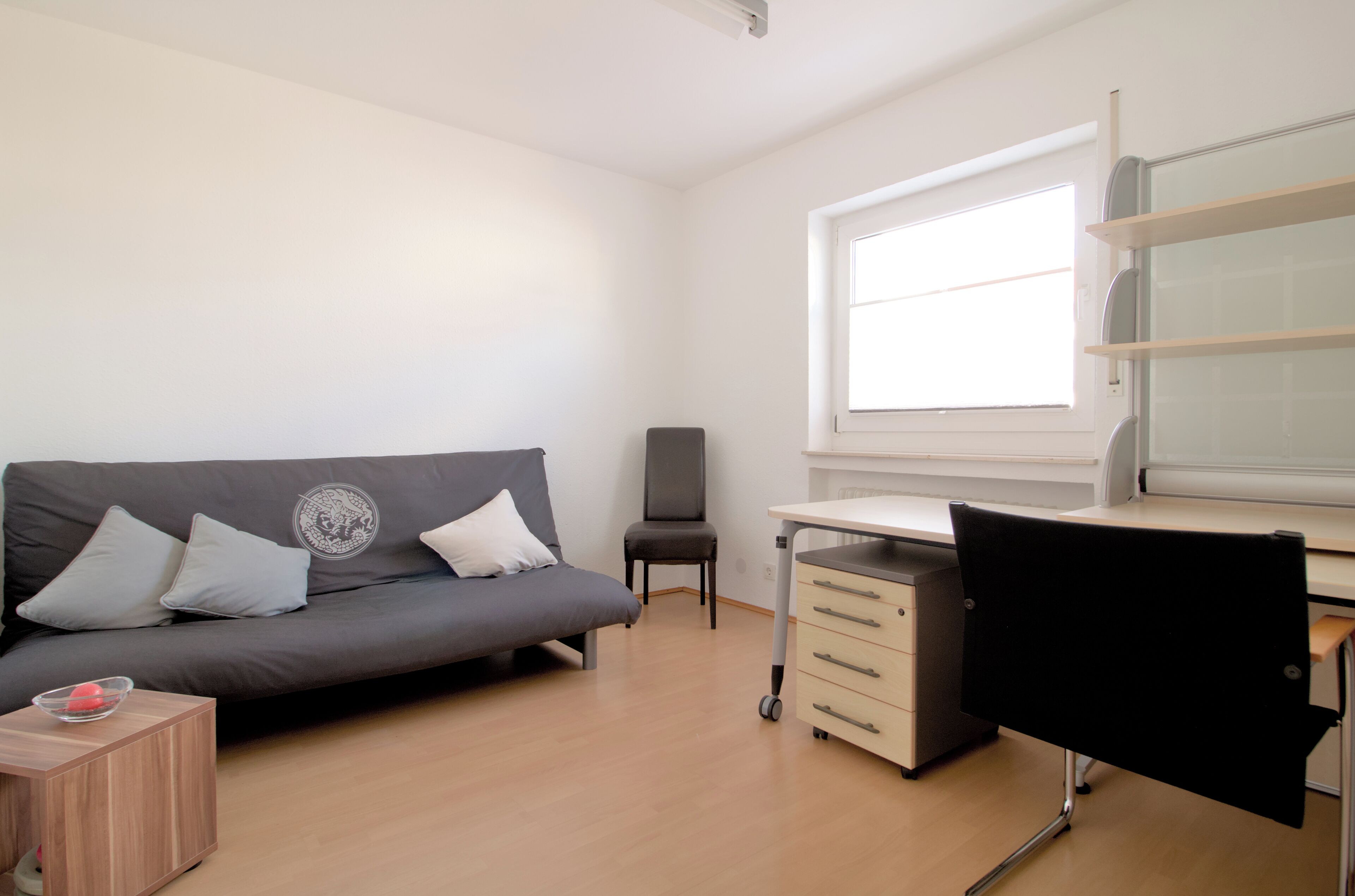A room with a grey sofa, a black chair, a desk with a computer, and a window.