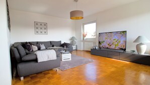 Ein modernes Wohnzimmer mit grauem Sofa, Holzboden, Flachbildfernseher und großem Fenster.