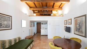 Een kamer met houten plafond, een eettafel met stoelen, een bank en een keuken in de achtergrond.