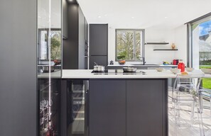 A modern kitchen with a large window, dark cabinetry, and a bar area with clear chairs. - -EDGE- Modern house with garden, near beach. (Névez)