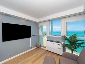 A modern living room with a flat-screen TV, large windows with a view of the sea, and a ceiling fan. - NEW! Steps to the Beach, Pool, AC, View! Close to Attractions (Luquillo)