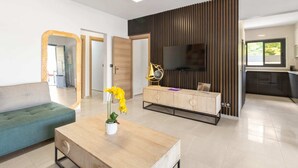 A modern living room with a sofa, a wooden coffee table with a vase of flowers, a TV mounted on a wooden panel wall, and a kitchen area in the background. - Résidence Dorée - Seven Bedroom Villa, Sleeps 14 (Sainte-Anne)