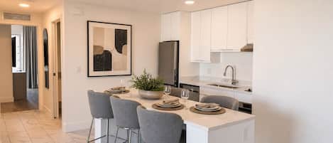 A modern kitchen with white cabinets, a central island with bar stools, and a dining area with a plant centerpiece.