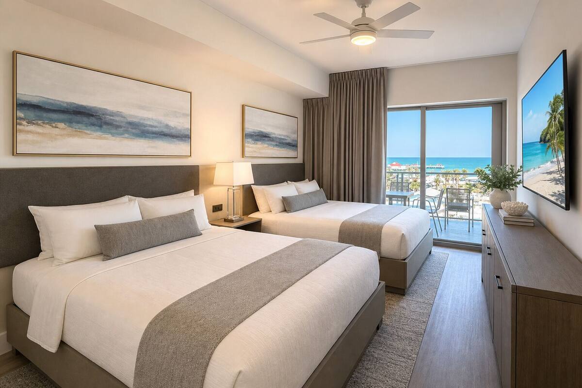 Habitación de hotel con dos camas, ventilador de techo, ventana grande con vista a la playa y obras de arte en las paredes.