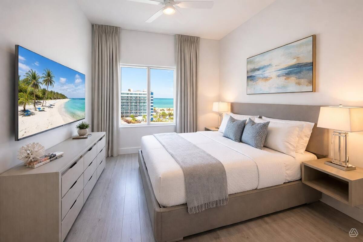 Habitación de hotel con una cama grande, un televisor mostrando una escena de playa, un cuadro y vista a un edificio y playa a través de la ventana.
