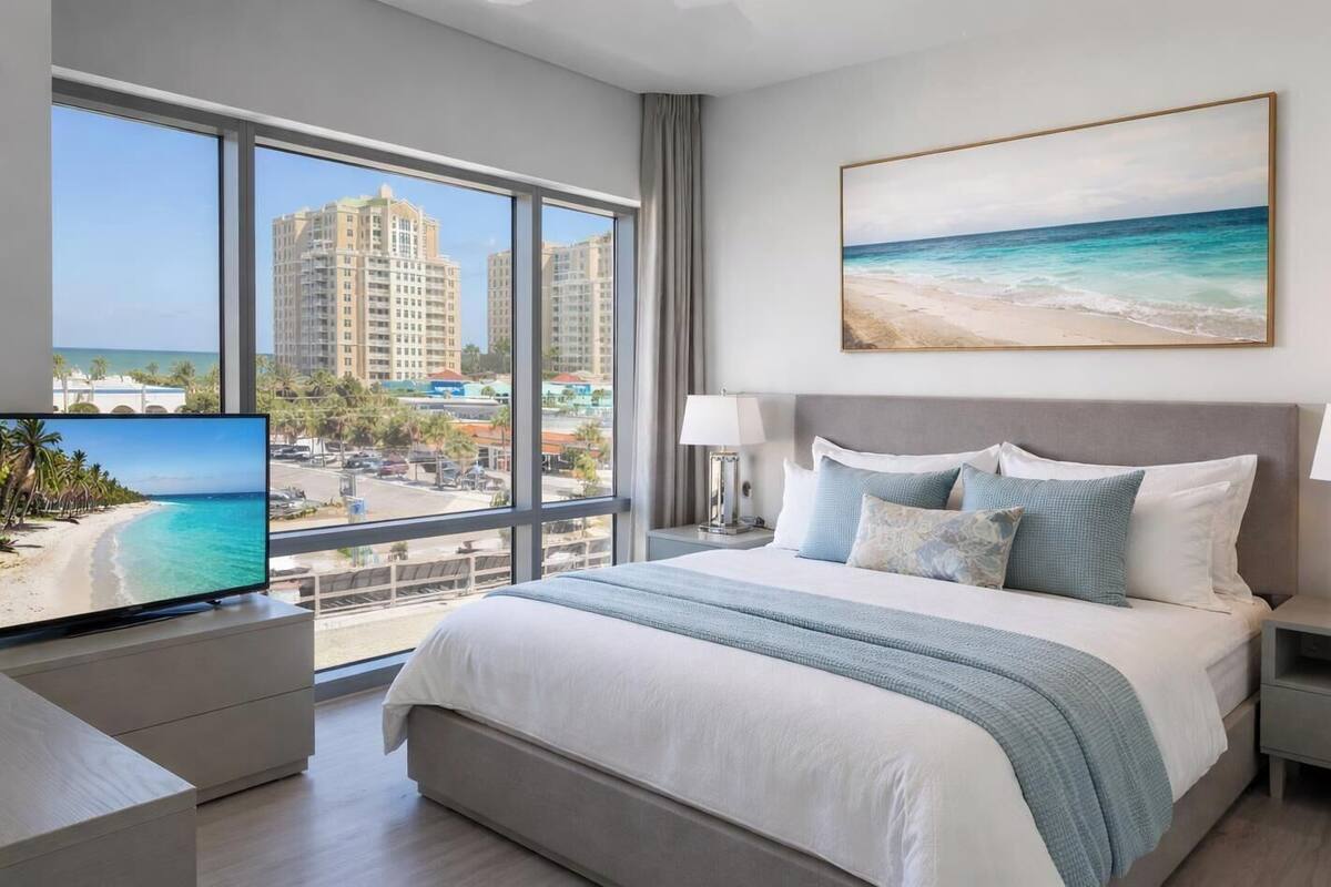 Un cuarto de hotel con una cama grande, un televisor sobre un mueble, un cuadro de una playa y vistas a edificios altos y una playa a través de la ventana.