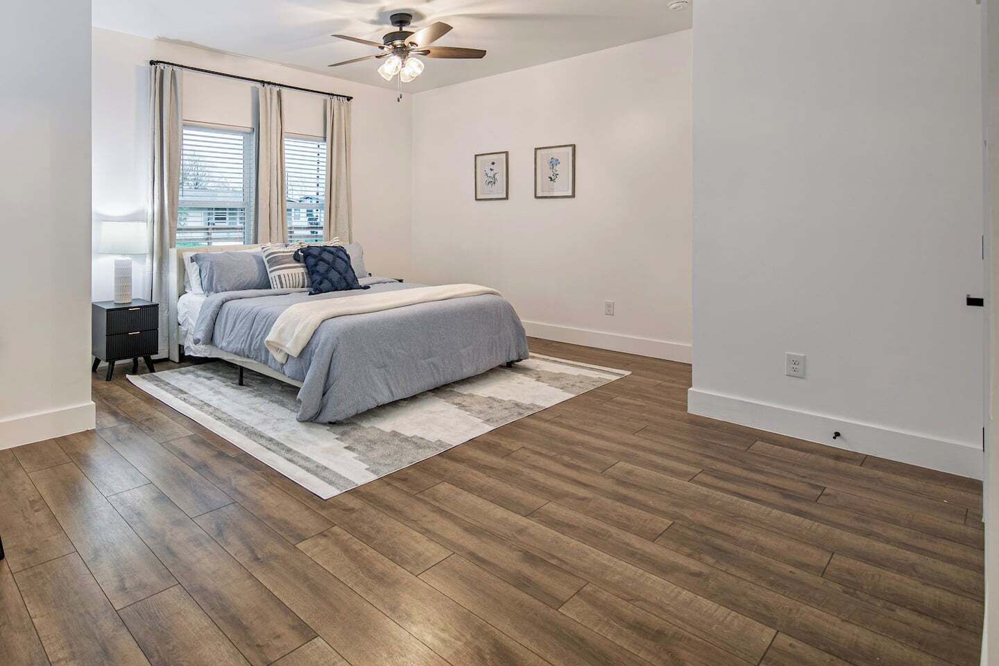 A bedroom with a bed, a nightstand, a ceiling fan, a window with blinds, and a wall with two framed pictures.
