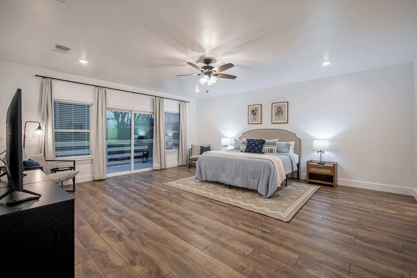 Une chambre à coucher comprenant un lit, un bureau, une télévision, un ventilateur de plafond et une grande fenêtre avec des stores.