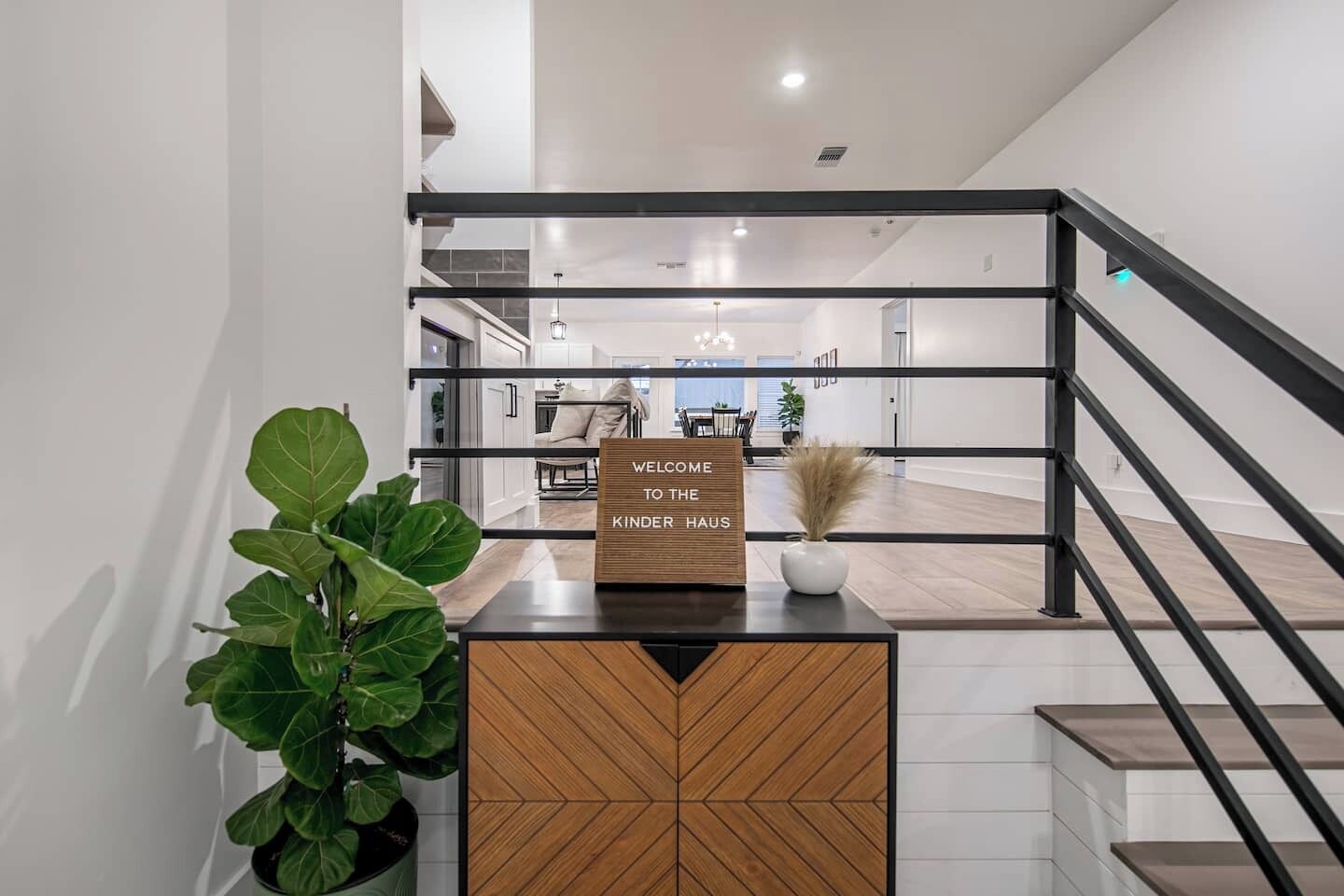 Un intérieur moderne avec un comptoir d’accueil en bois, une enseigne indiquant « BIENVENUE À LA KINDER HAUS », et un escalier avec des rampes noires.