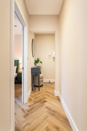 A hallway with a herringbone wooden floor, a small table with a plant, and a door leading to a room with a bed and a lamp. - The Brighton Bliss - Sleeps 7 (Brighton)