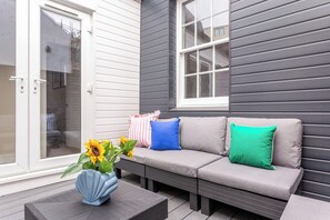 A gray outdoor seating area with a sofa and cushions, a blue vase with sunflowers, and a door with a glass panel. - The Brighton Bliss - Sleeps 7 (Brighton)