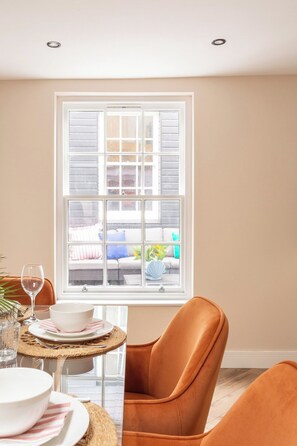 A dining area with a table set for two, a window with blinds, and a view of a balcony. - The Brighton Bliss - Sleeps 7 (Brighton)