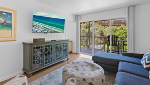 Ein Wohnzimmer mit einem großen Fernseher, einer Anrichte, einer Couch und Blick auf einen Balkon mit Gartenmöbeln.