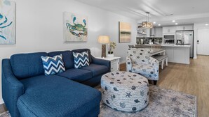 Ein modernes Wohnzimmer mit einem blauen Sofa, einem Hocker und passenden Sesseln, ein Essbereich mit einer Bank und eine Küche mit weißen Schränken und Geräten.