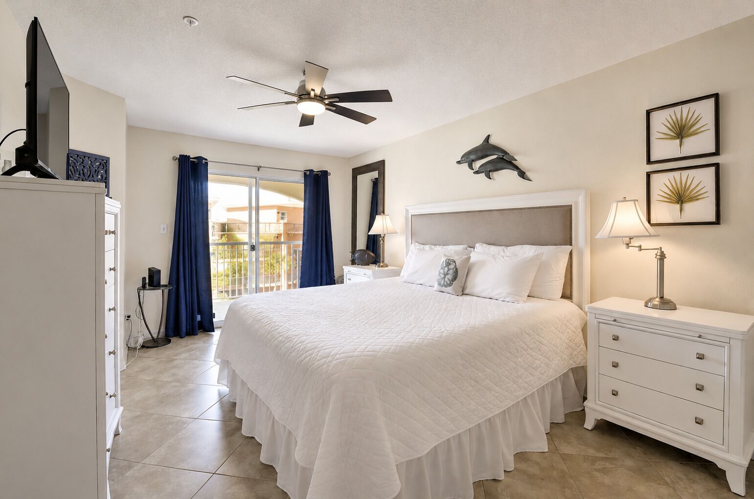 A hotel room with a bed, a nightstand, a lamp, a ceiling fan, a balcony with curtains, and a wall with dolphin artwork.