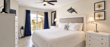 A hotel room with a bed, a nightstand, a lamp, a ceiling fan, a balcony with curtains, and a wall with dolphin artwork.
