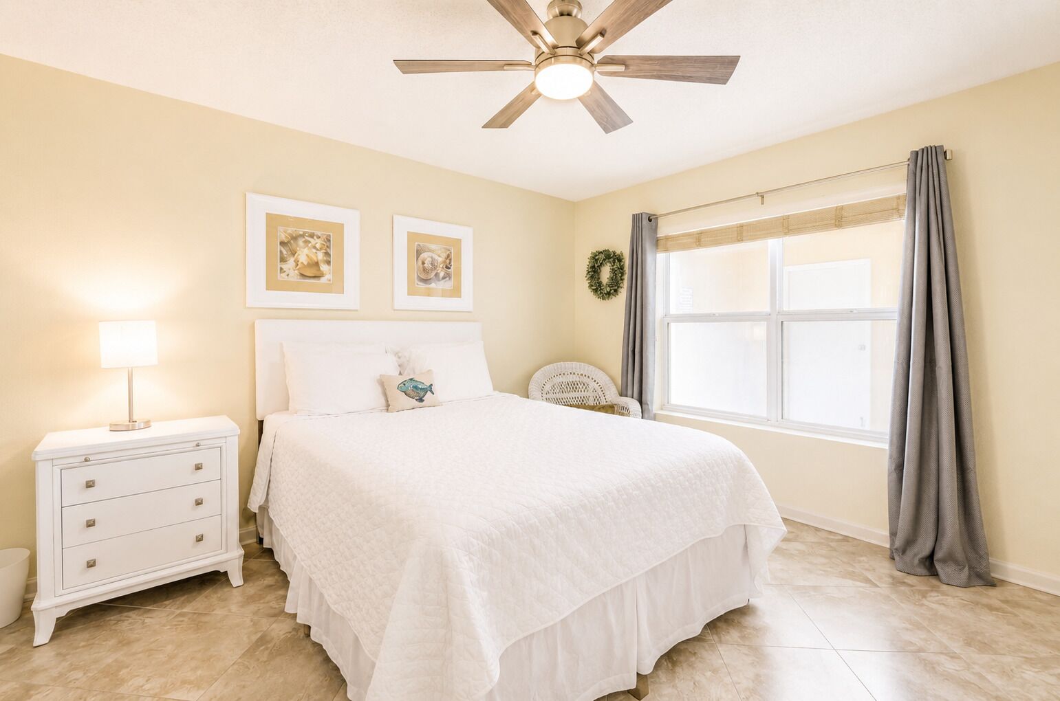 A bedroom with a bed, nightstand, lamp, two framed pictures, a ceiling fan, a window with curtains, and a wreath.