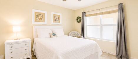 A bedroom with a bed, nightstand, lamp, two framed pictures, a ceiling fan, a window with curtains, and a wreath.