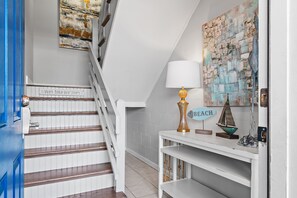 A staircase with a sign that reads 'Wipe Your Flip Flops,' a white console table with a lamp, and a wall with a nautical-themed painting.
