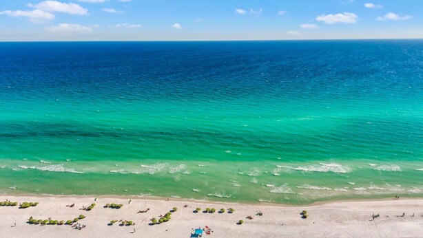 Ein Strand mit klarem türkisblauem Wasser, einem sandigen Ufer und einigen Sonnenschirmen.