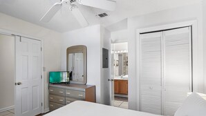 Ein Schlafzimmer mit Bett, Schminktisch, Fernseher, Deckenventilator und einem durch eine offene Tür einsehbaren Badezimmer.