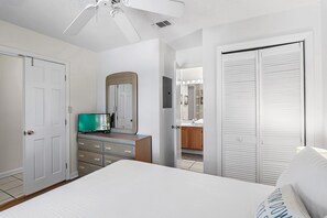 Ein Schlafzimmer mit Bett, Schminktisch, Fernseher, Deckenventilator und einem durch eine offene Tür einsehbaren Badezimmer.
