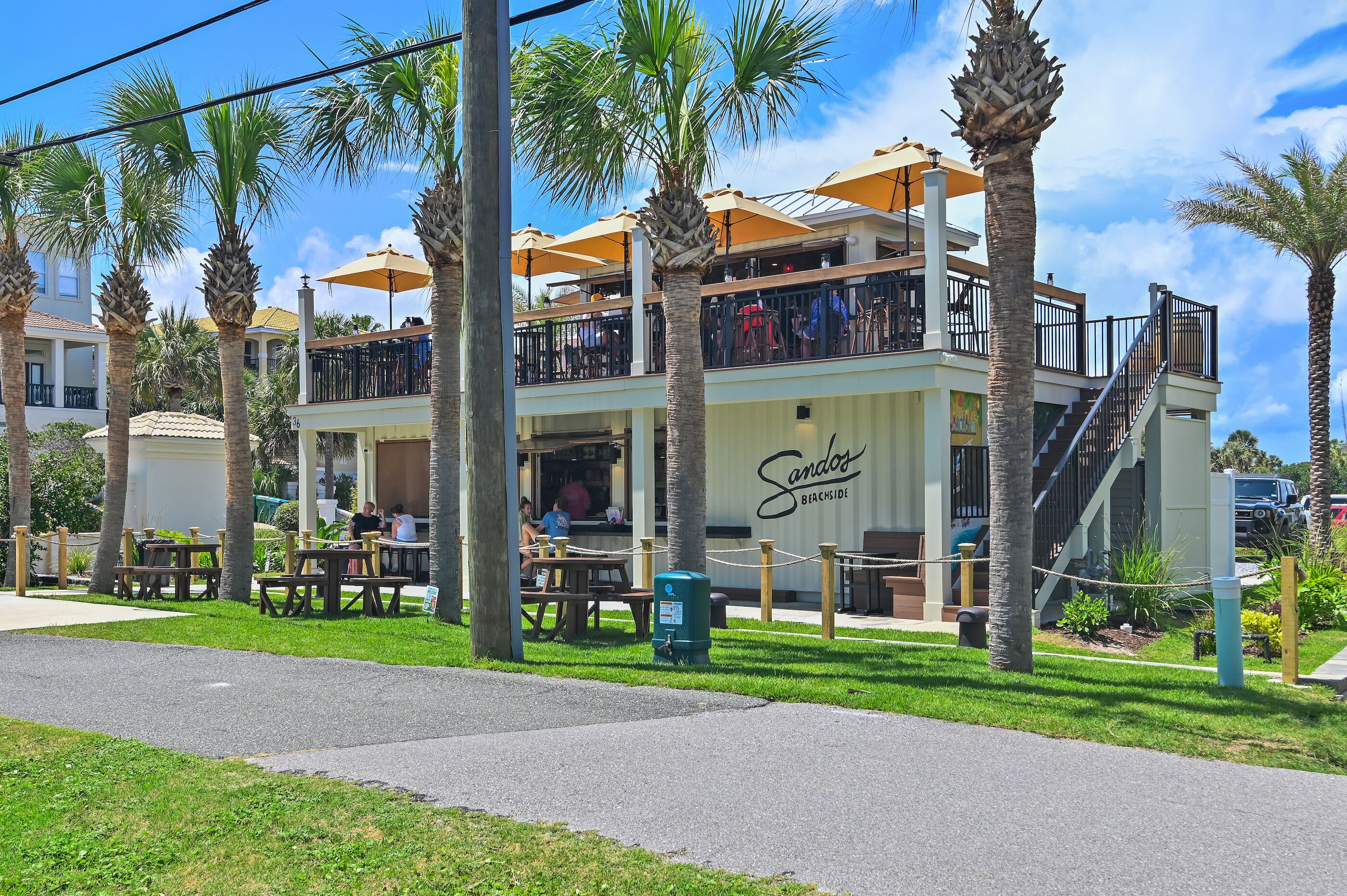 A two-story building with outdoor seating, palm trees, and a sign reading 'Sandoz Beachside.'