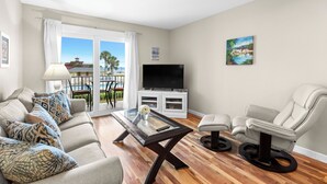 Un soggiorno con un divano, un tavolino in vetro, una TV su un mobile, una poltrona e un balcone con vista su una piscina e degli edifici.