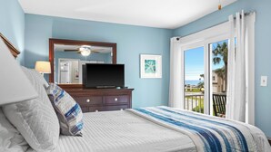 Una camera da letto con un letto, una cassettiera, una televisione, una ventola a soffitto e un balcone con vista sulle palme.