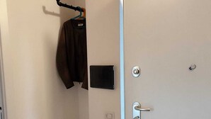 A room with a white door, a small white table, a coat hanger with a jacket, a black object on the wall, and a fire extinguisher. - Thumbelina House (Sapri)