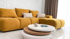 Un divano color senape con numerosi cuscini, un tavolino da caffè bianco con un oggetto decorativo e un pavimento in marmo.