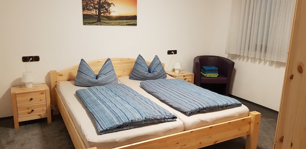 A wooden bed with two pillows and a blanket, a nightstand with a lamp, a chair, and a wall-mounted power outlet.