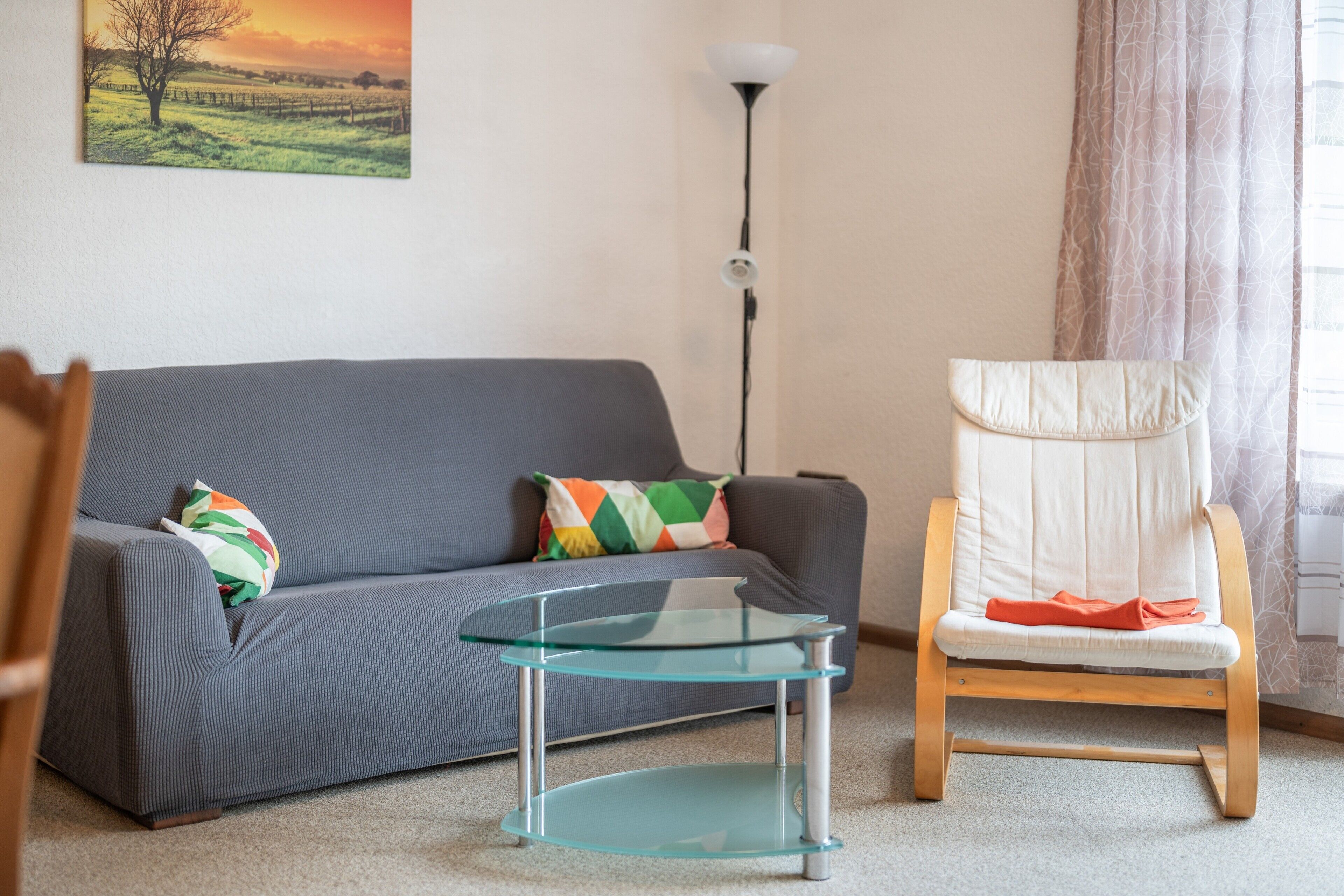 Ein Wohnzimmer mit grauem Sofa, einem Glastisch, einem Holzstuhl und einer Stehlampe.
