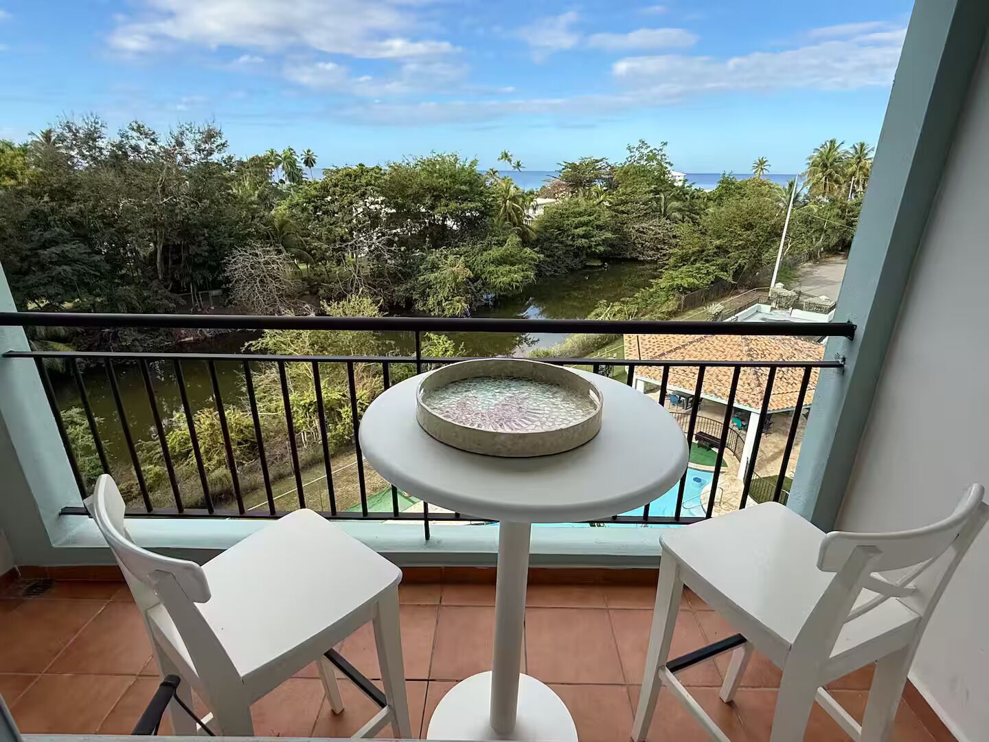 Een balkon met een ronde tafel en twee stoelen, met uitzicht op een zwembad en weelderige begroeiing.