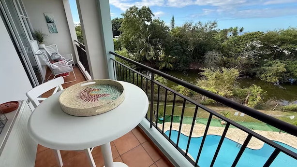 Un balcón con una mesa blanca, sillas y un plato decorativo. Se ve una piscina y abundante vegetación.