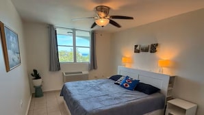 Un dormitorio con cama, ventilador de techo, lámparas de noche, una ventana con cortinas y una mesita de noche.