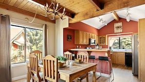 Un intérieur rustique avec une table à manger en bois, des chaises, un lustre et une cuisine dotée de meubles en bois et d’une fenêtre.
