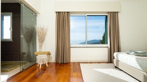 Ein Schlafzimmer mit großem Fenster, einem Bett, einem kleinen Hocker und einer Glasdusche.