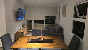 Un comedor moderno con una mesa de madera, sillas tapizadas de gris, un frutero, un televisor montado en la pared y un sofá al fondo.