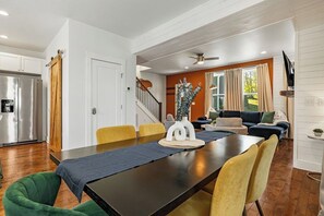 Eine moderne Küche mit einem Edelstahlkühlschrank, ein Essbereich mit einem dunklen Holztisch und gelben Stühlen sowie ein Wohnzimmer mit einer Couch und einem großen Fenster.