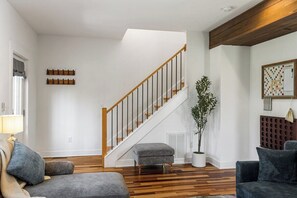 Ein modernes Wohnzimmer mit einer Holztreppe, einem grauen Sofa, einem kleinen Hocker, einer Topfpflanze und einem Scrabble-Spiel auf der Wand.