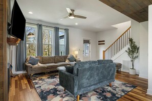 A living room with a sofa, a TV mounted on the wall, a ceiling fan, and a staircase. - Blue Ridge House with Hot Tub - 204 (Asheville)
